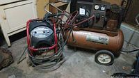 Photo of a twin cylinder air compressor on wheels alongside a red frame Powermate gas pressure washer with multiple hoses, all placed on a concrete floor.