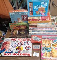 Photo showing assorted vintage children's crafts and board games stacked, including Yarn Scribbles, Touch 'n Tuck, Rafia Basket, Beadcraft Outfit, Pot Holders and various board games with colorful illustrated boxes.