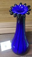 Tall cobalt blue blown glass vase with black vertical stripes and ruffled black-trimmed edge, displayed empty against wood backdrop.