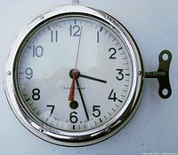 Front view of the closed clock showing white dial with black numbers, red second hand, and winding key attached.