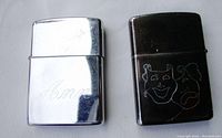Two Zippo lighters shown, one polished silver 'Ann' engraved, other dark with comedy/tragedy mask engraving.