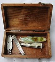 Open wooden box containing three small pocket knives and one mother-of-pearl nail file showing compact size and handles