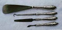 Four antique sterling silver women's beauty tools arranged horizontally on a flat surface: shoehorn, boot lace hook, cuticle trimmer, nail file.