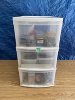 Three-drawer clear white plastic organizer box on wood surface, filled with various small containers and bags of hardware items.