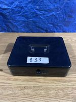 Closed black metal cash box with silver handle on top and combination dial lock on the front, placed on a wooden surface against a blue backdrop.