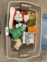 Top view of a plastic bin containing various Christmas decorations such as plush ornaments, ceramic cookie jars (gingerbread man, Santa Claus), a white stocking with reindeer decoration, and a star ornament.
