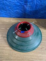 Green metal Christmas tree stand holder with red mounting area and adjustable eye bolts for tree trunk securing.