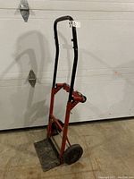 Side angle view of manual hand dolly showing red and black metal frame, black handle, two wheels, and metal base plate.