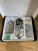 Open box showing white handheld device with control buttons, LCD screen, and attached electrode pads with wires. Includes two gray adhesive electrode pads, and one wire set in tray.
