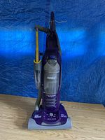 Full front view of purple upright vacuum showing the overall body, dust bin, handle, and attached hose with yellow Hair Raiser brush.