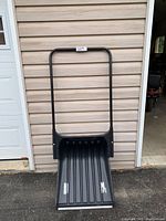 Front view of the large black snow pusher showing the blade and the handle against a tan siding wall.