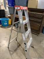 Photo of the Lite step ladder showing its two-sided aluminum frame, orange top cap with brand name, and rubber feet.