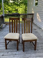 Two wooden dining chairs shown outdoors on a wooden deck. Chairs have vertical slat backs and beige upholstered seats.