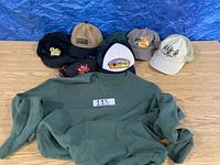 Lot of six assorted baseball caps and one green crewneck sweater laid on floor.
