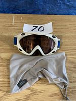 Top view of white Carrera ski goggles with blue strap resting on wooden surface alongside a grey protective pouch.
