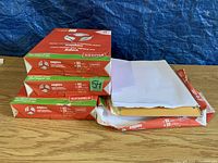 Three unopened reams of Staples printer paper stacked with one unopened ream beside several loose sheets of white paper on a wooden surface against a blue tarp background.