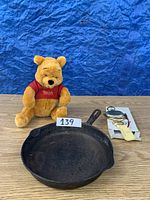 Winnie The Pooh plush, cast iron pan, and brass door knocker in original packaging on a wood surface with blue backdrop.