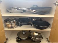 Photo of three shelves inside a cabinet showing various cooking pans and pots neatly arranged including frying pans, saucepans, steamer insert, colander and double grill pan with glass lid.