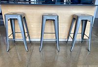Front view of the three silver metal bar stools showing square seats with handle cut-outs and four legs with crossbars.
