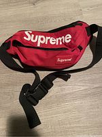 Red Supreme fanny pack with white logo and black adjustable strap laid flat on light wood floor.