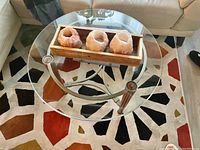 Top and angle view of a round glass coffee table with three metal legs and ornate salt candle holders in wooden tray on top.