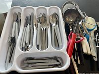 Photo showing a white plastic cutlery tray filled with assorted cutlery and extra kitchen utensils alongside.
