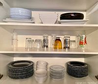 Shelves with white dishes and assorted glassware including clear glasses and a brown glass, plus black and white speckled plates and multiple white bowls and small plates.