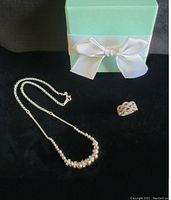 Sterling silver necklace with polished silver balls, ring with sapphires, and green box with white bow on black cloth.