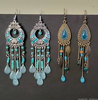 Photo showing two pairs of long drop earrings: one silver-tone with turquoise beads and teardrop shapes, and one brass-tone with blue and orange beads and dangling chains