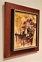 Framed oil painting depicting boats and buildings in earthy tones with red roof highlights, signed lower left.