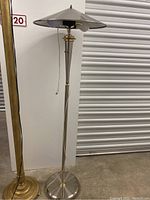 Full view of stainless steel and brass two-bulb floor lamp showing conical shade, slender stem, and round base with power cord visible on floor.