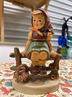 Front view of the Hummel figurine showing the girl sitting on a rustic wooden fence with a basket beside her, revealing details and paint quality.