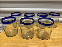 Photo showing 7 clear handblown glass tumblers with thick blue rims, round, sturdy shape