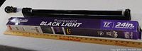 Fluorescent black light tube alongside its original purple and black branded box, with a measuring stick displaying approximate size.