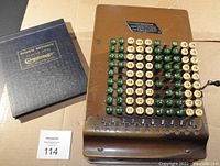 Top view of brass comptometer showing six columns of green and cream keys with numbers, alongside original instruction book in blue hardcover.
