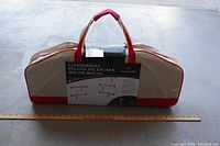 Full view of the unopened ladder ball game carrying case with a measuring stick for scale. Beige canvas with red handles and base with label attached.