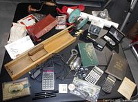 Overview of lot items including wooden grater, calculators, red push toy handset, metal spoons, decorative boxes, and paper ephemera displayed on table.