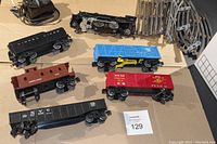 Photo showing six train cars including black steam locomotive chassis, coal car, flatcar with crane, boxcar, gondola and caboose with assorted curved metal track pieces and power supply unit visible