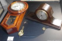 Two antique wooden wind-up clocks side by side on black surface. Left is Regulator wall clock with door and pendulum, right is Gilbert mantel clock with curved body. One key laid between them.