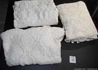Three hand-crocheted white tablecloths folded and displayed on a black surface, showing varying crochet patterns and sizes.