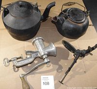 Photo showing two cast iron kettles, manual meat grinder, and a cherry pitter on cardboard.