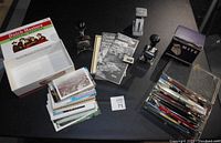 Overview of lot showing box of Dutch Masters on left, multiple office stampers, vintage booklets, and a stack of postcards, with clear plastic box of pens and pencils on right.