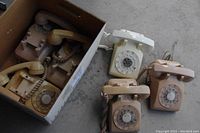 Overall view of five rotary dial telephones arranged on floor and in box showing three beige/pinkish phones outside and two inside box.