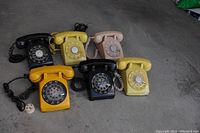 Front-side view of six vintage rotary dial telephones in black, yellow, beige, and orange colors arranged in two rows on concrete floor.