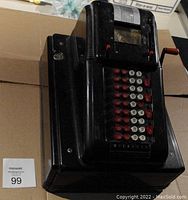 Front angle view of the McCaskey manual adding machine highlighting red and white keys and wooden crank handle.