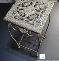 Full view of the metal plant stand showing the patterned square top and curved legs on a dark background.