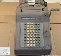 Front view showing gray metal body, multiple keys, numeric display, and crank handle.