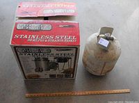 Photo of the original packaging box for the Oklahoma Joe Classic Stainless Steel 30 Qt Turkey Fryer alongside a propane tank showing scale with a wooden ruler.