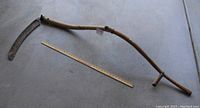 Full length image of antique wooden handled scythe with attached rusted metal curved blade, placed on a concrete floor with measuring ruler for scale.