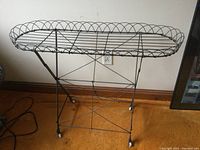 Full front view of antique wire plant stand showing oval top, wire frame, cross-bracing, and ceramic wheels.